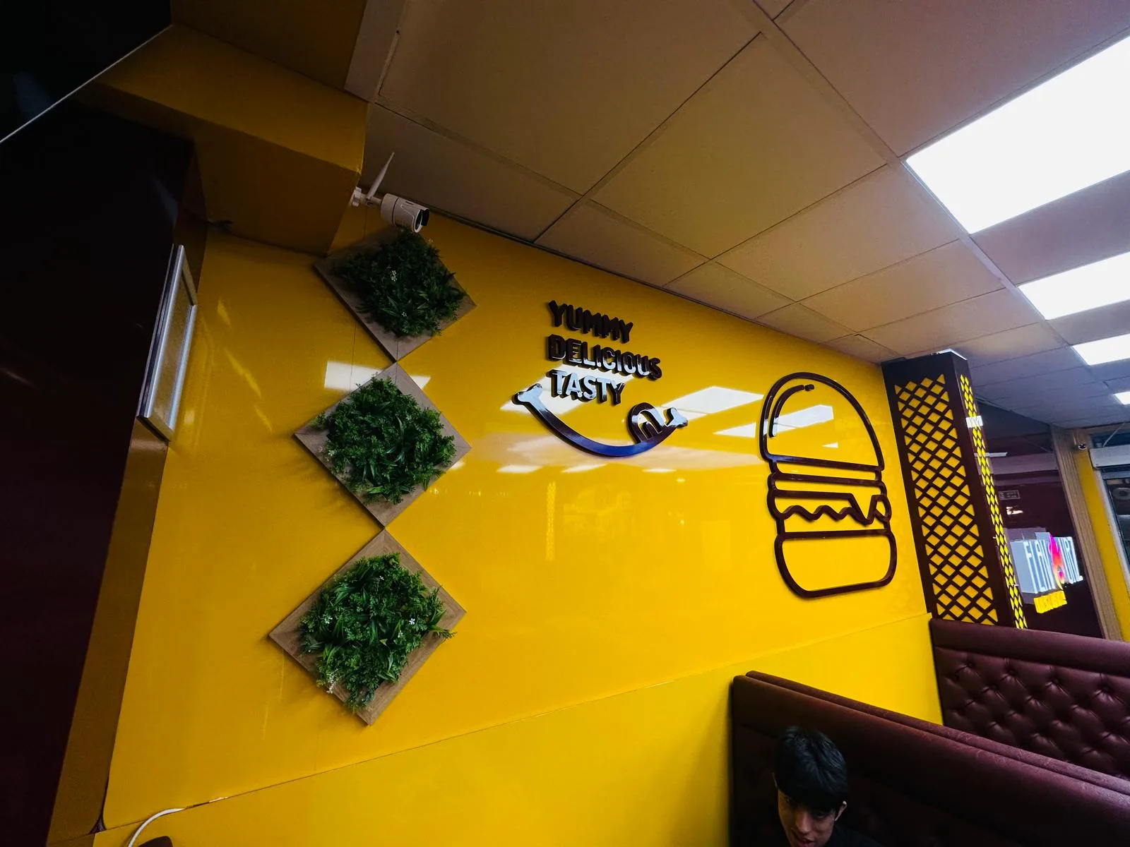 Interior wall branding with dark 3D lettering and a burger graphic on a glossy yellow wall with green foliage panels