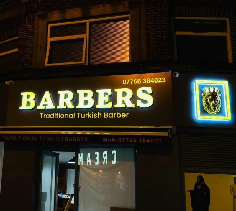 Illuminated shop sign with glowing yellow lettering on a lit fascia and a blue LED sign on a barber shop front at night