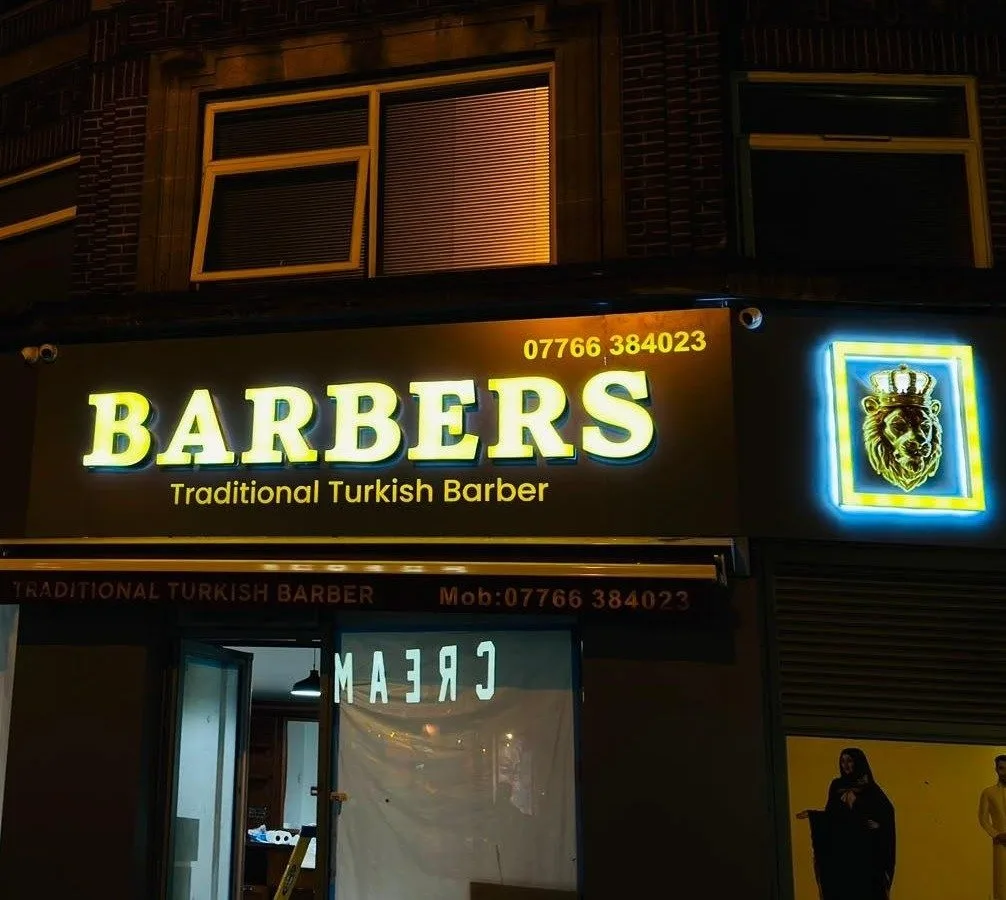 Illuminated shop sign with glowing yellow lettering on a lit fascia and a blue LED sign on a barber shop front at night